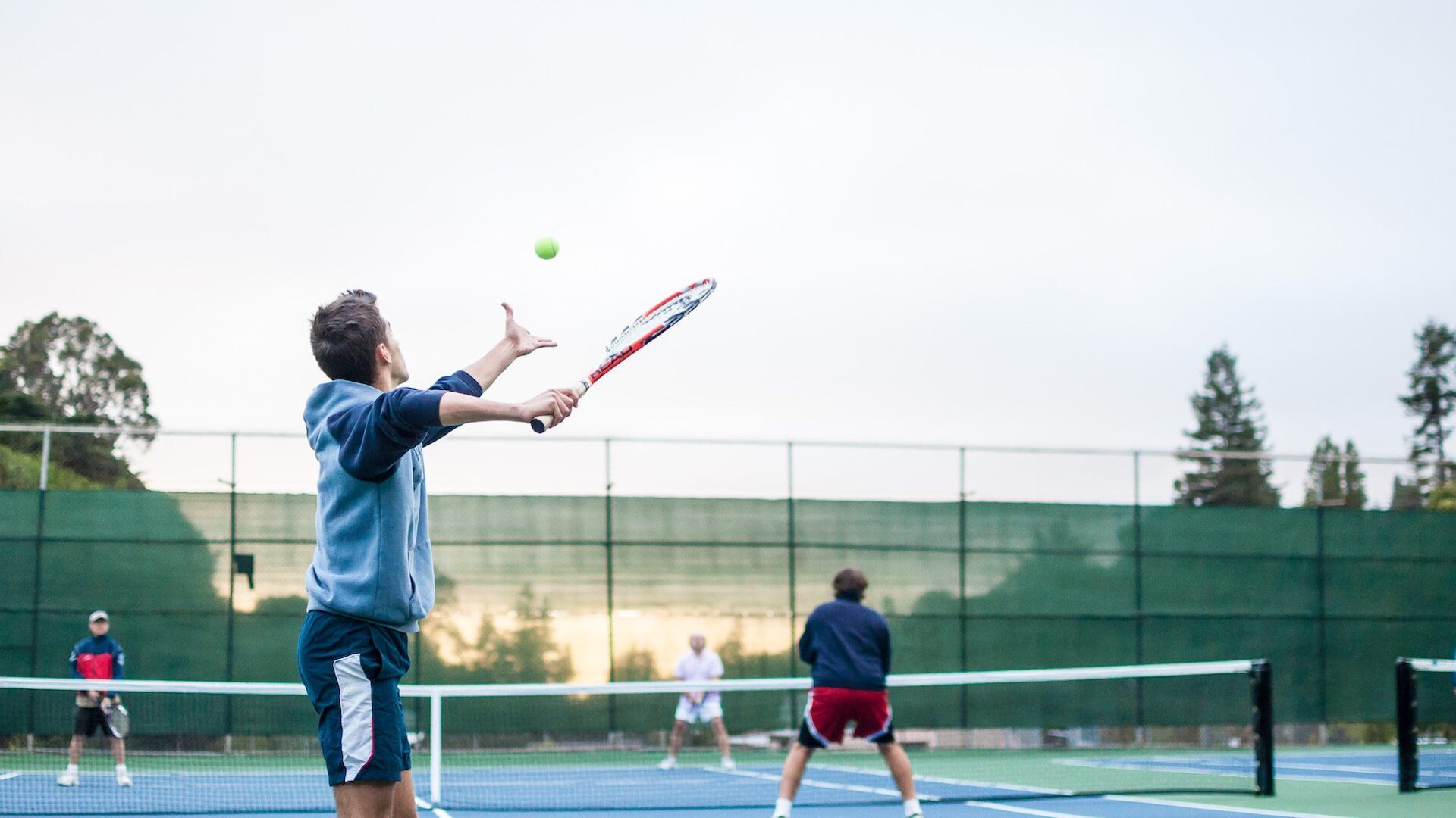 Group of amateur tennis players on a tennis court, man in the foreground is tossing a ball up in preparation of serving