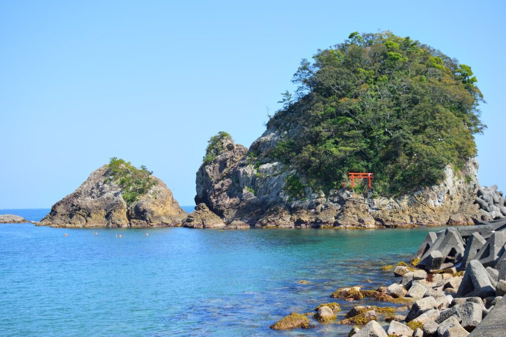 A bushy island sits in the background with a red torii gate sitcking out, while in the foreground peaceful clear waters sit with rocks poking out - covered in moss.