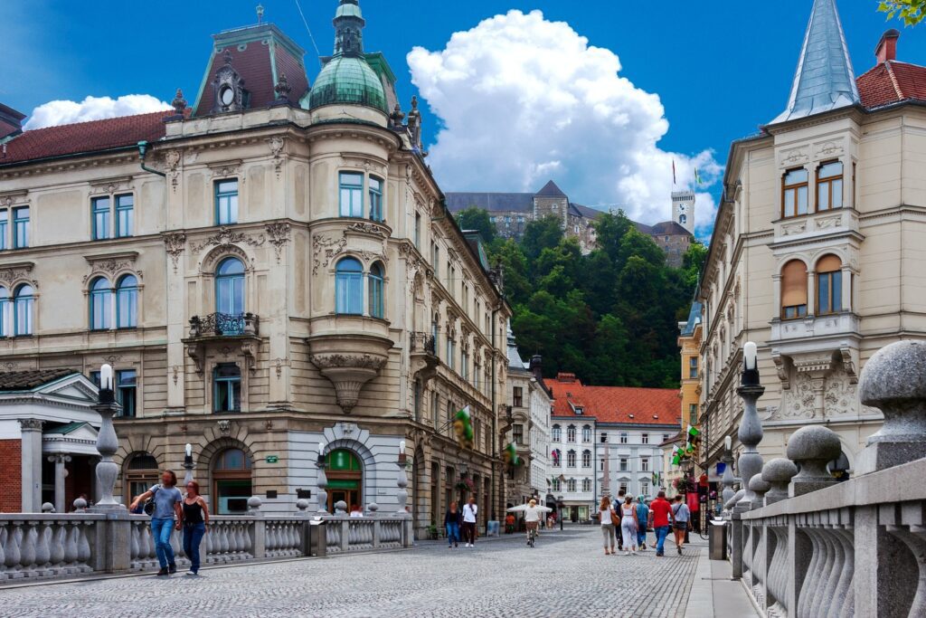 A lively street scene in a family-friendly European city with ornate buildings and a castle on a hill in the background, under a cloudy sky.