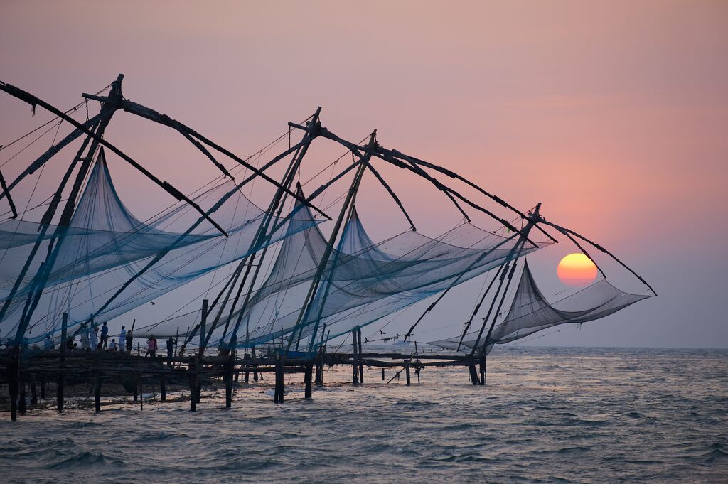 The sun sets in Kerala. The ocean stretches out into the horizon and the sun is orange and red. A pier filled with sails and nets take up the foreground