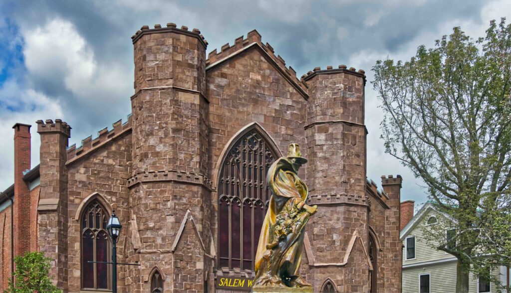 Close-up of a building in Salem which is a witch museum. In front of the building is a gold statue. This is one of the best places to visit for Halloween