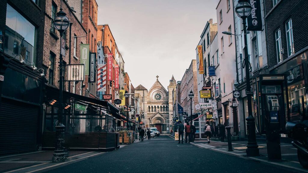 A high street in Dublin, Ireland. Signs of the stores are on the outsides of the buildings, with a church at the back and in the center.