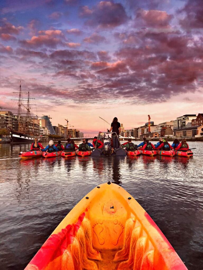 POV view of sitting in a kayak in Dublin on the river, with a horizontal row of other kayakers sitting in front. The sky is crimson and the city looks gorgeous