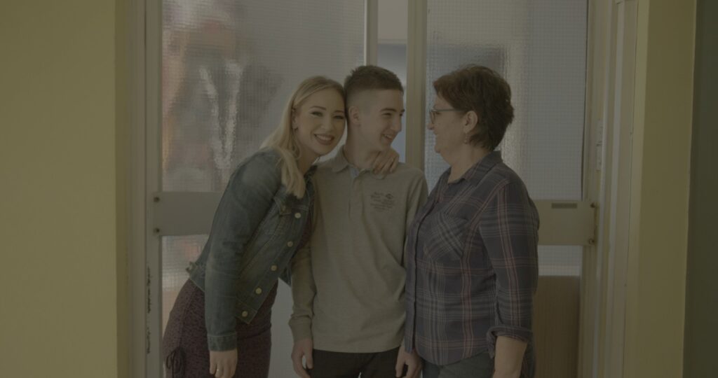 Jasmina, Senada and Aldin stand close together smiling in the entrance to their home of Sarajevo, Bosnia