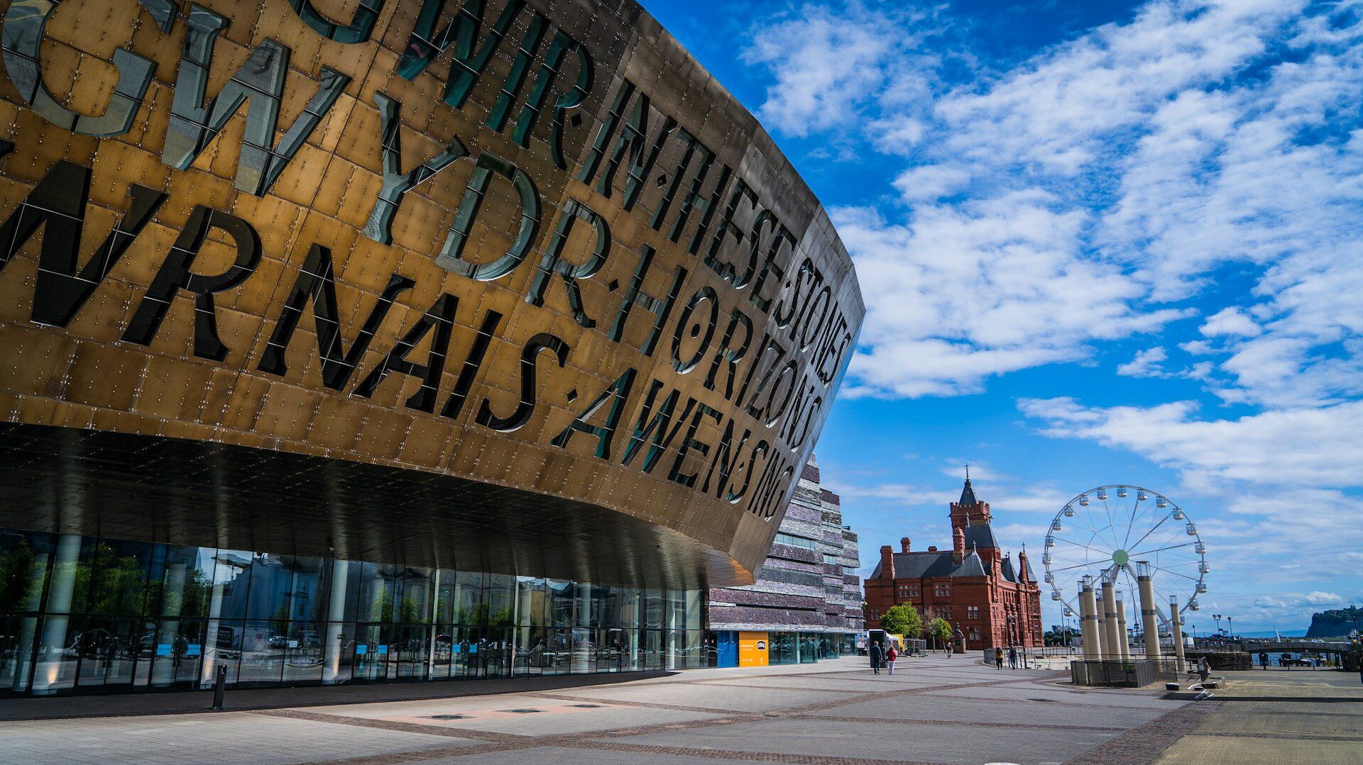 Photo of a large modern building in the foreground, with a metal facade stencilled with Welsh text. In the background, a large redbrick building and a ferris wheel