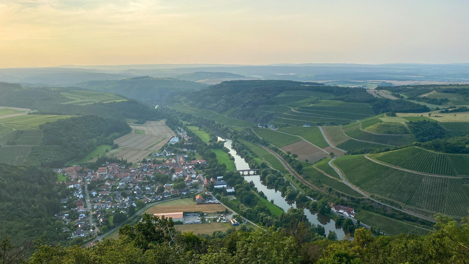 Aerial view of the Rhine Valley. We can see the river running past a town, with a arched bridge crossing it. We can see crop fields and hills stretching into the distance