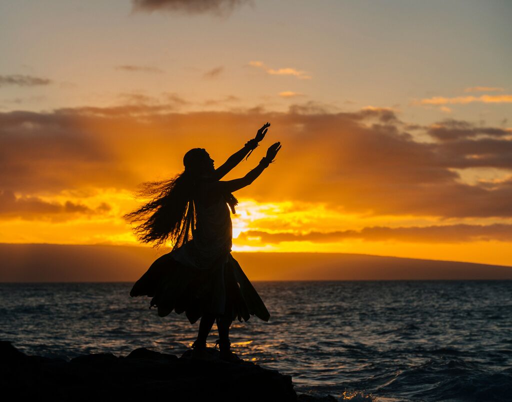 Woman with hulu attire on silhouetted during the sunset, dancing and looking out at sea