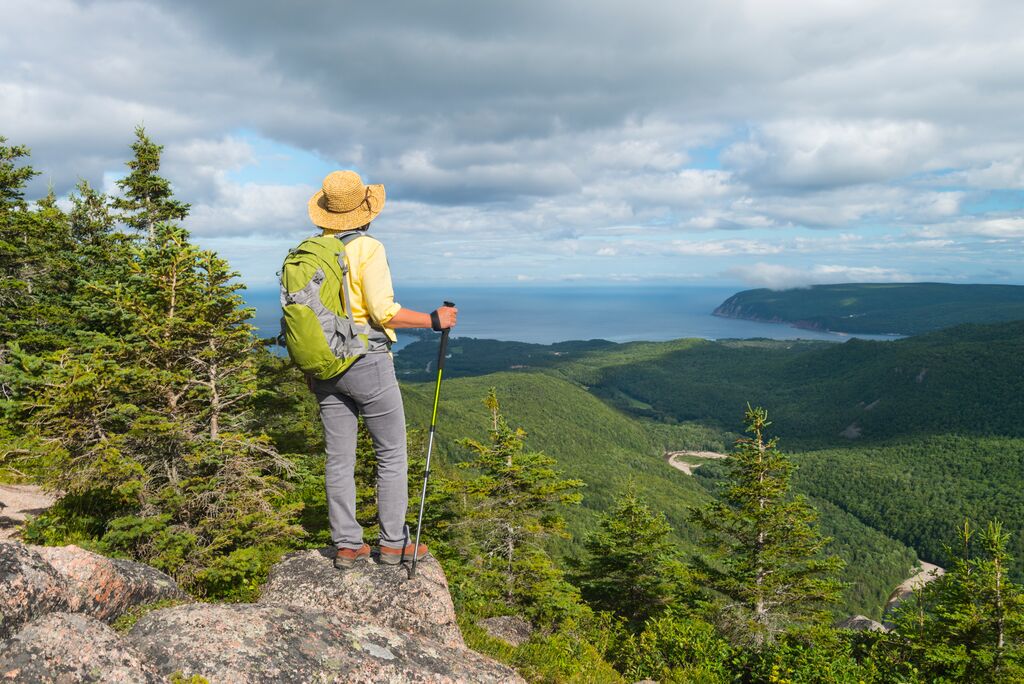 A photo of a mature woman from behind wearing a hiking backpack and a straw hat, holding hiking poles, standing on a rocky outcrop overlooking densely forested terrain