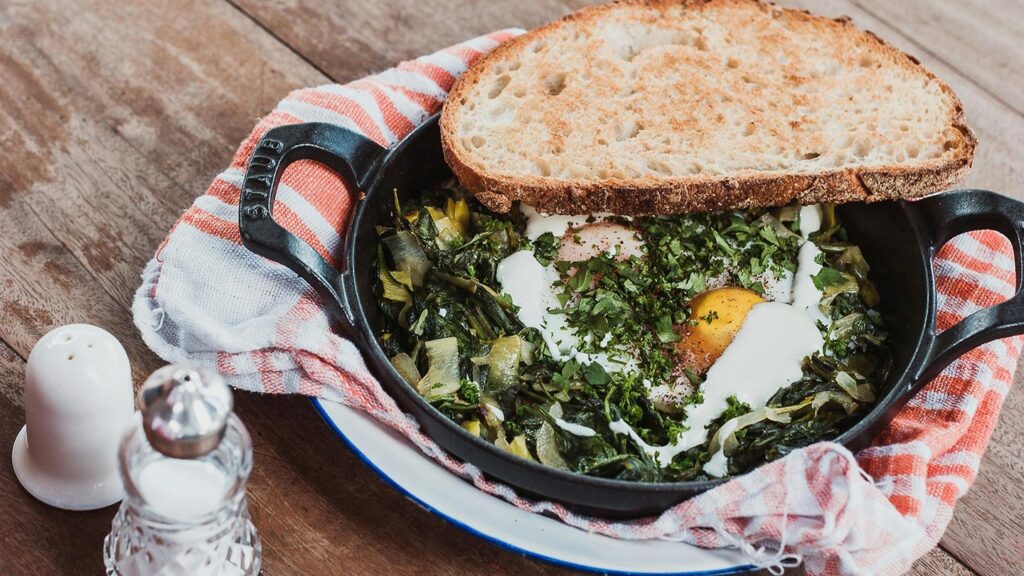 A photo of a cast iron skillet containing sauteed green vegetables and eggs, sitting on a red and white checked cloth