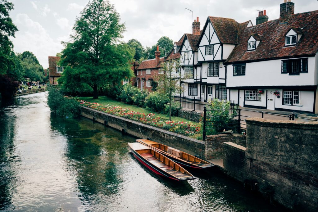 Two small wooden row boats sit along a quiet canal outside of a hotel in Canterbury, England.