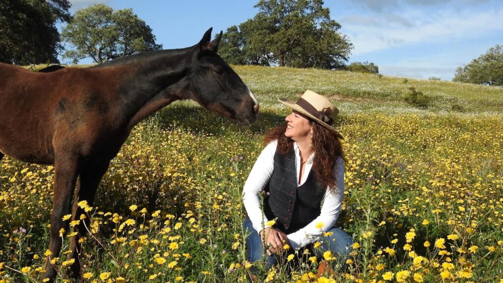 A woman and a horse smiling at each other while sat in a field of yellow flowers