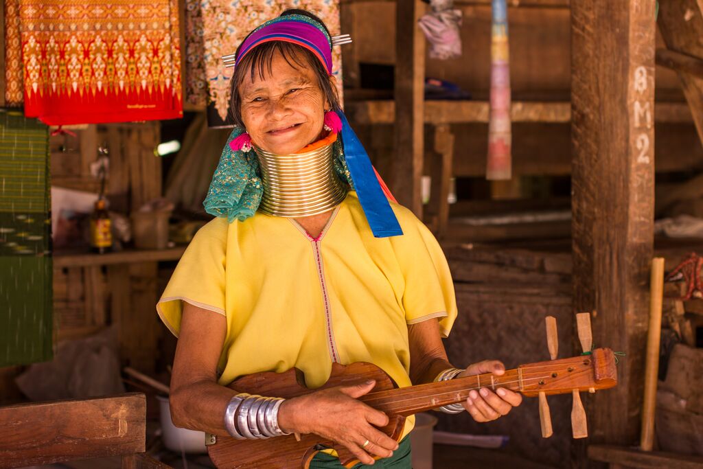 A tribal woman with a traditional neck ringlet smiling while playing a small guitar.