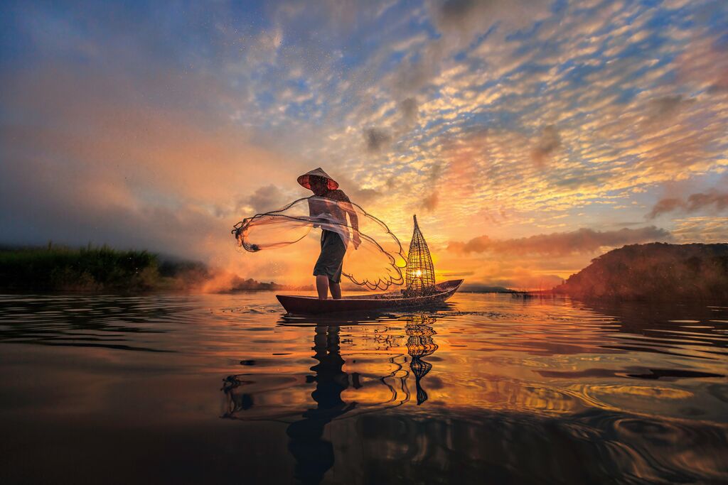 A fisherman casting a net while wearing a straw hat, standing in the middle of a body of water on a canoe.