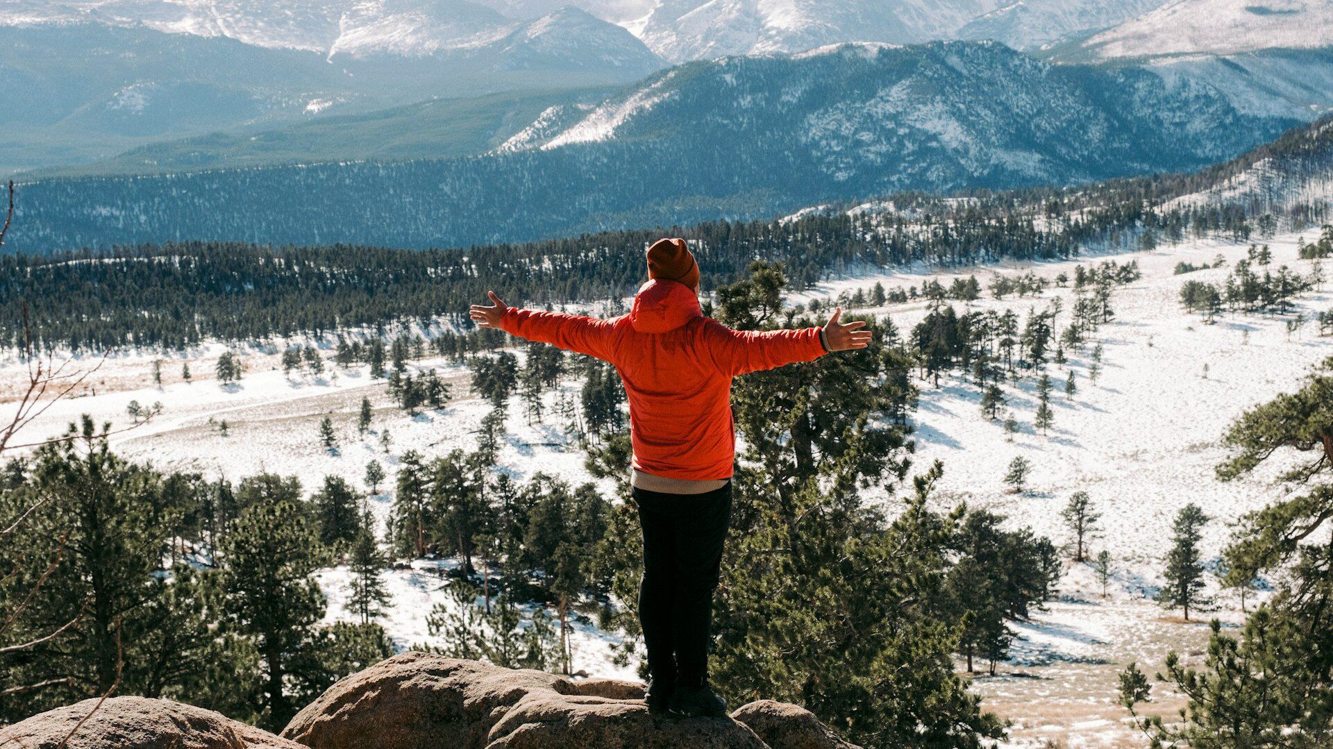 Man in a red coat standing with arms outstretched looking over a snowy mountainous landscape