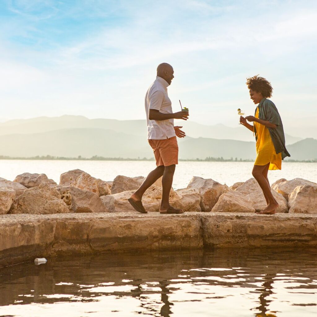 Two travelers holding cocktails are about to embrace while walking on a small path lined with rocks, sea and mountains in the background.