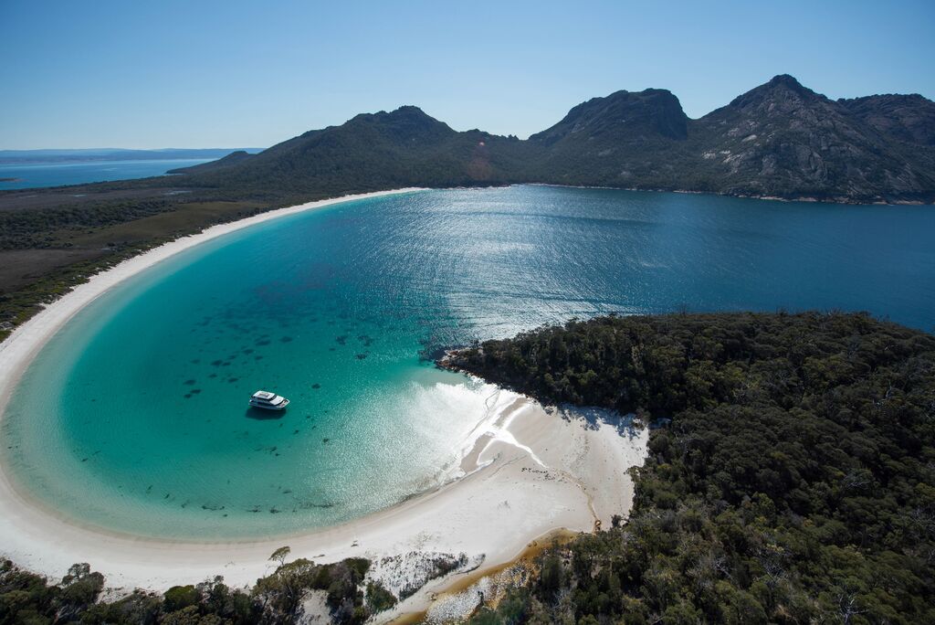 A birds-eye view of Wineglass Bay, in Tasmania. Turquoise waters are surrounded by lush vegetation and mountains. A lone ship sits in the bay.