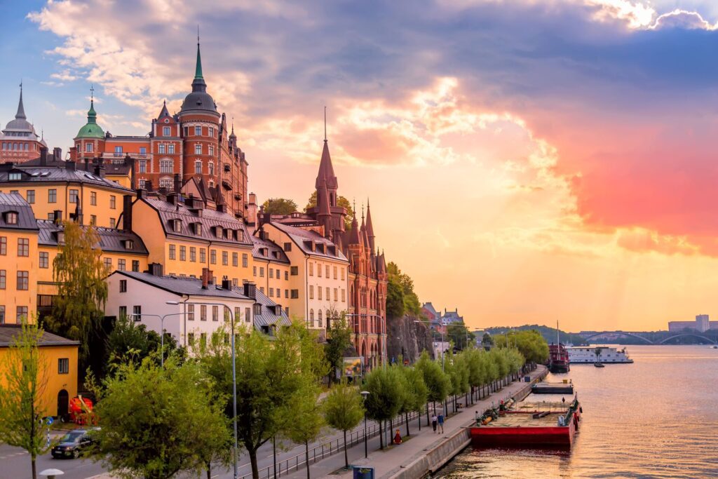 Riverside in Stockholm. Multicolored buildings look out over the river, trees line the promenade, and a violet colour fills the sunset sky.