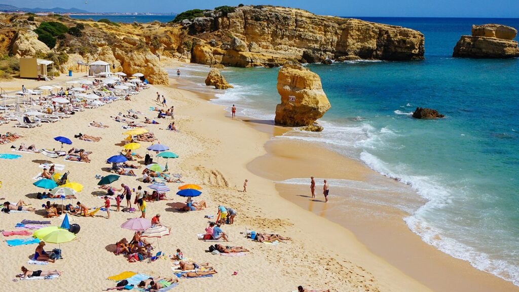 A crowded sandy beach with colorful umbrellas and people enjoying the sun, surrounded by rocky cliffs and the blue sea, exemplifies why travel to Portugal is so appealing.