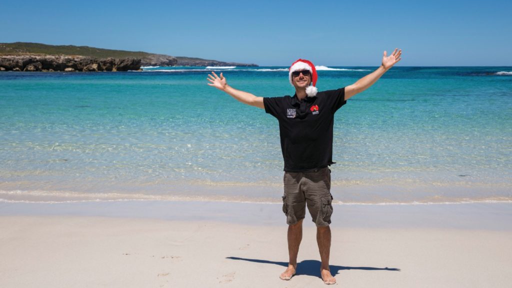 Man in santa hat and sunglasses standing on a sunny beach with arms outstretched, celebrating Christmas Down Under.