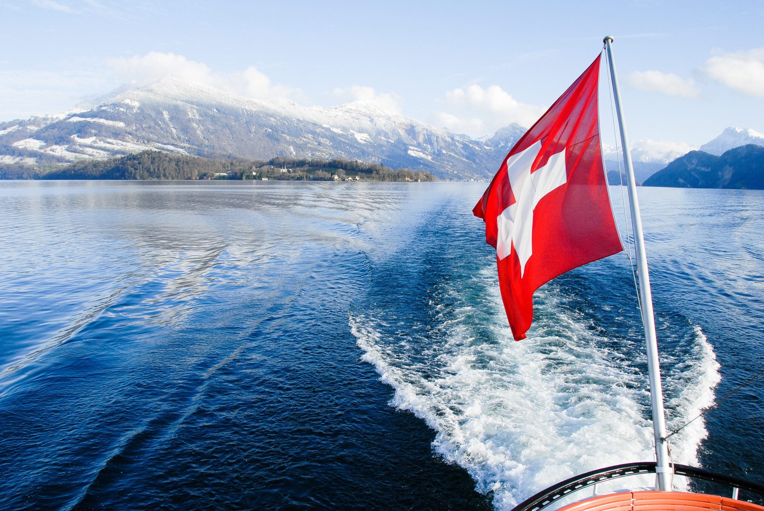 Welcome to Switzerland: Swiss flag waving on the stern of a boat moving through a lake with snow-capped mountains in the background.