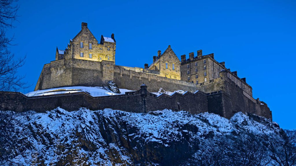 Edinburgh castle illuminated at dusk against a December twilight sky, with snow-covered cliffs in the foreground.