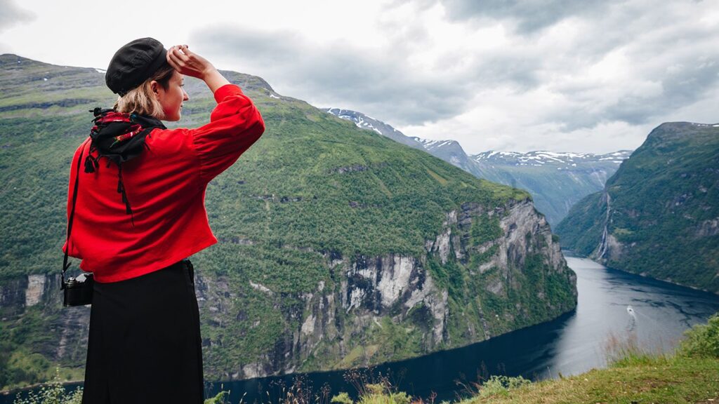 Woman in a red jacket looking at a scenic fjord surrounded by lush mountains on her romantic Europe tour.