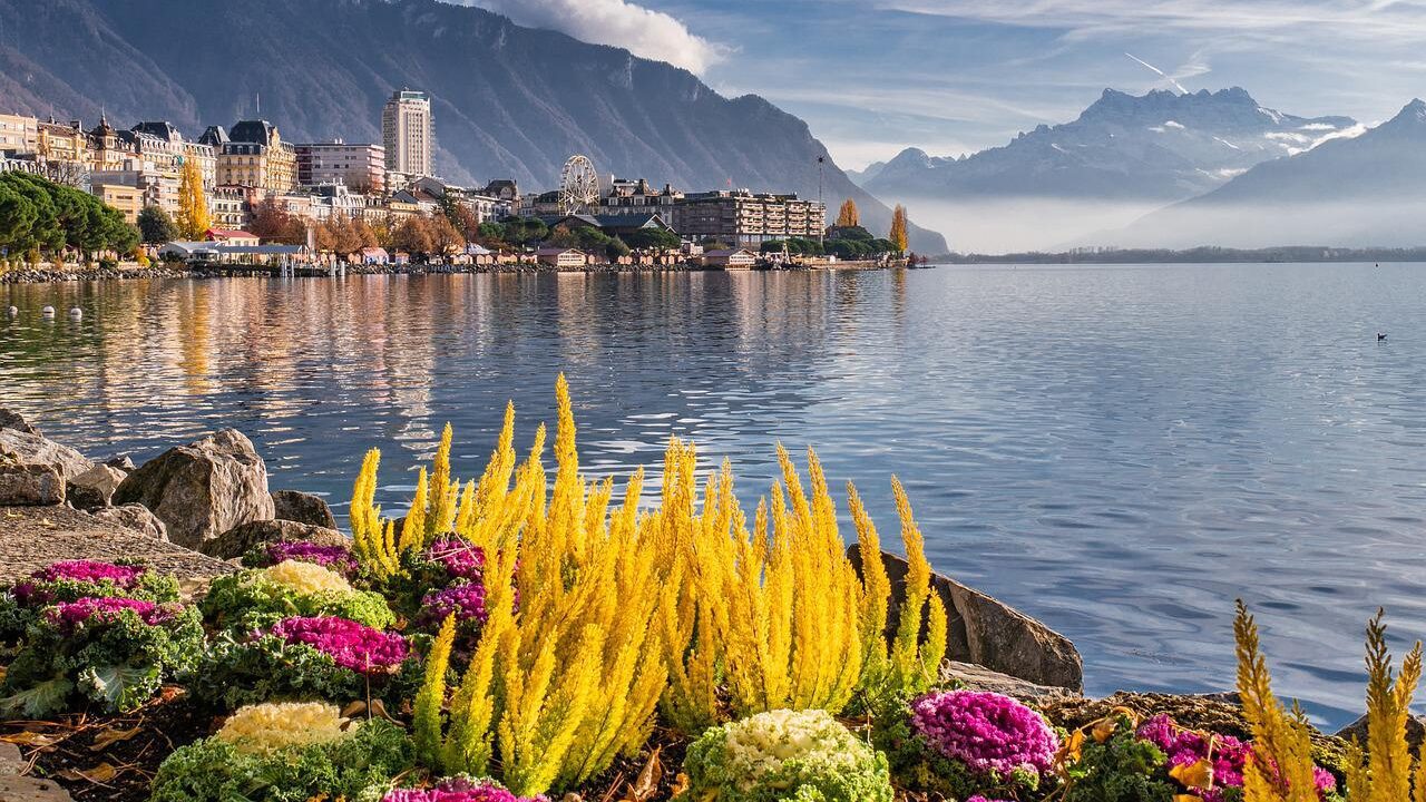 Scenic view of a lakeside town in Switzerland with colorful flowers in the foreground, calm water, and mountains under a clear sky.