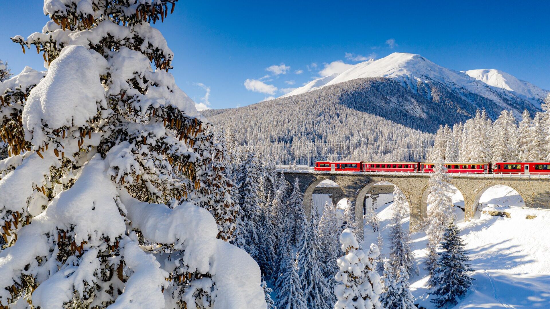 Red train travelling to Switzerland, crossing a snowy viaduct in a mountainous landscape with snow-covered trees and clear blue sky.