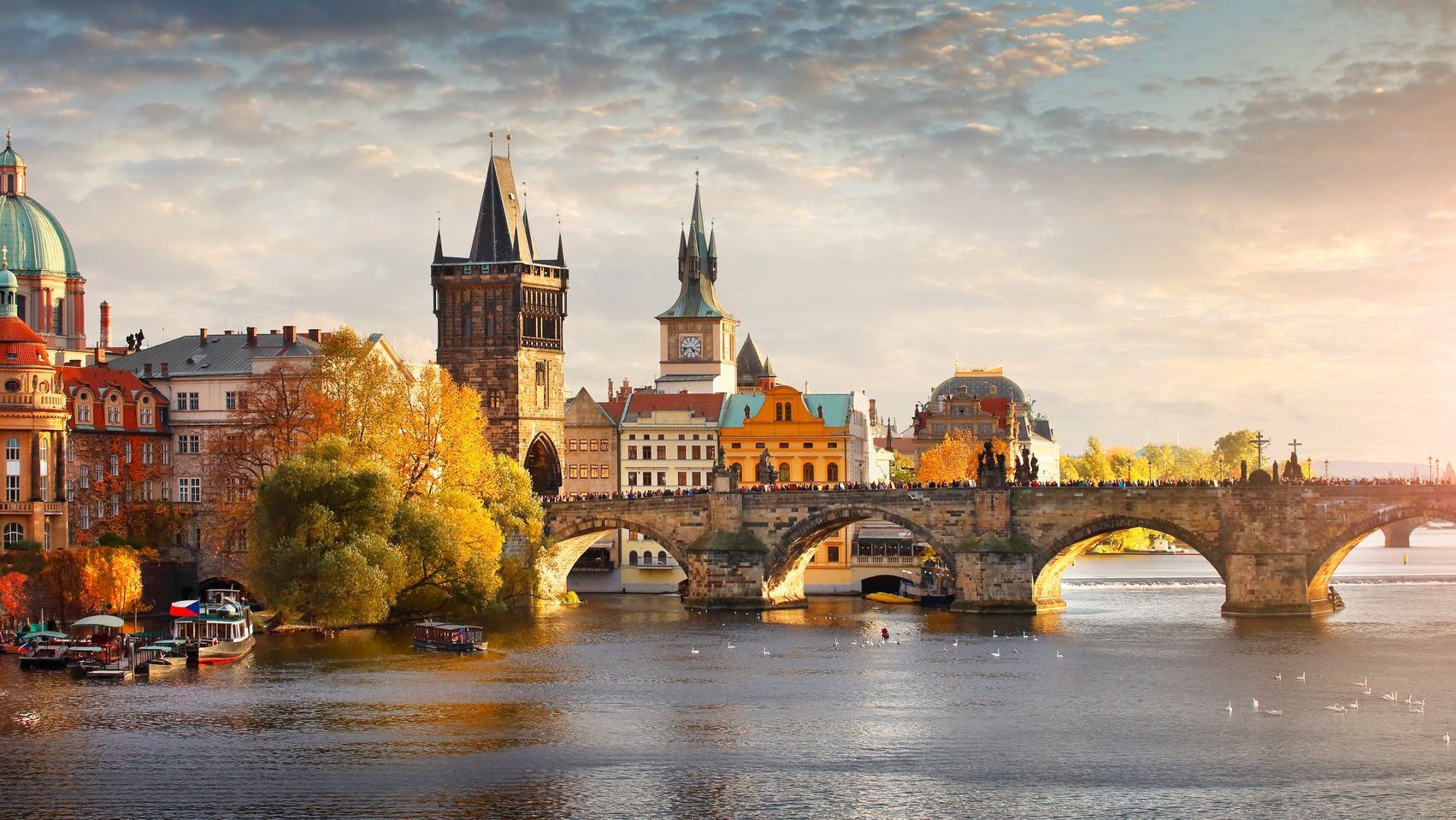 View of a bridge over a river with a historic town skyline on one side, with visible clock towers and church steeples
