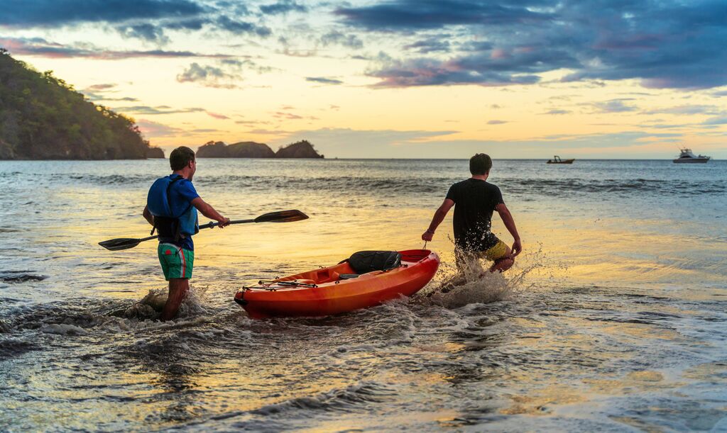 Two men walking a canoe out into the shallow sea off of a beach, one carry the paddle