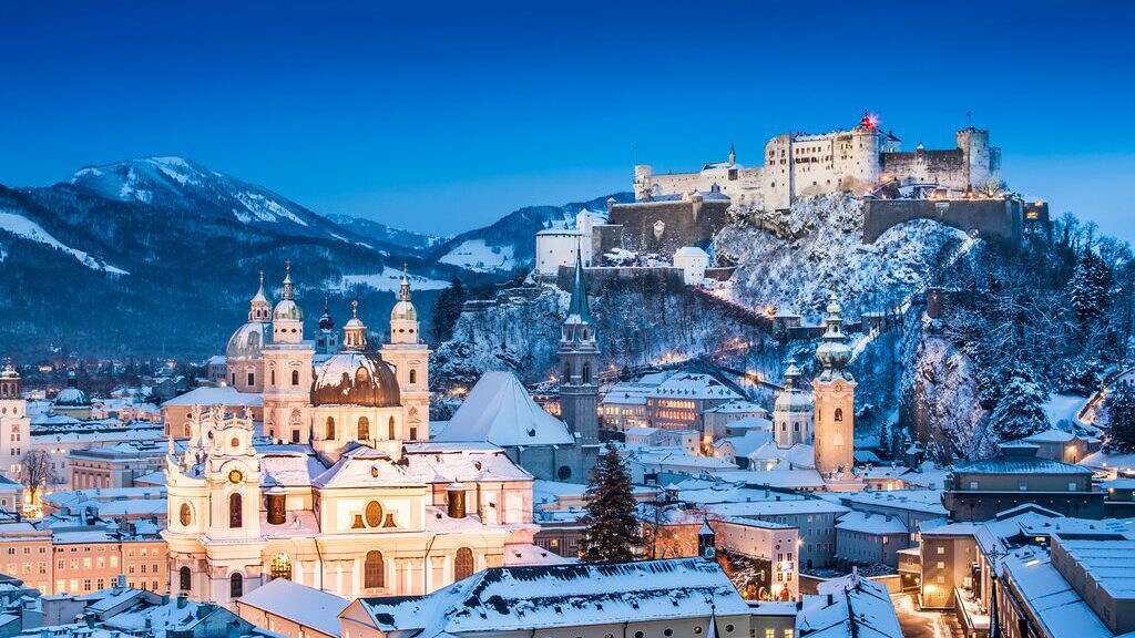 Winter wonderland view of Salzburg at dusk with Hohensalzburg Fortress lit up, and snow-covered roofs and mountains in the background.