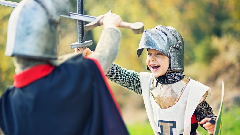A child in medieval knight costume play-fighting with an adult in a helmet, both holding swords in a sunny outdoor setting, perfect for family travel tips.
