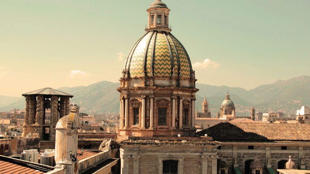 View of a cityscape featuring an ornate dome with a patterned roof, encountered on a walking tour, surrounded by other historic architectural elements, with mountains in the background.