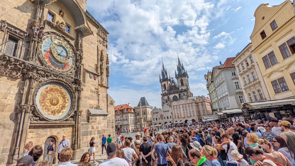 Crowded square in imperial Europe featuring the astronomical clock and the Church of Our Lady before Týn, under a blue sky.