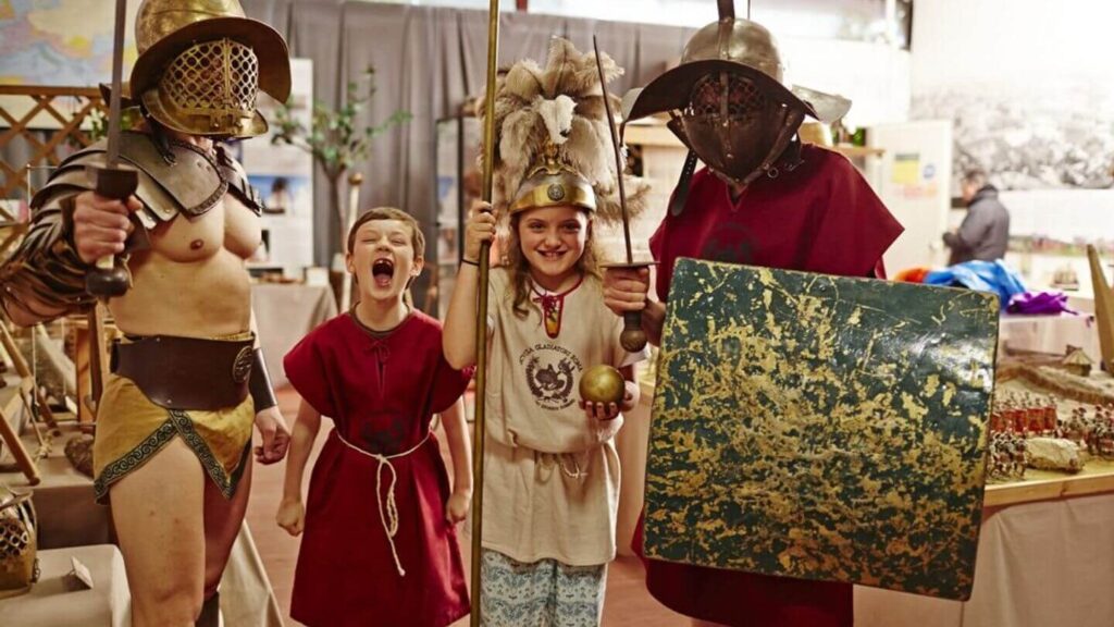 Two children in Roman-style clothes standing next to men in Roman-style gladiator outfits