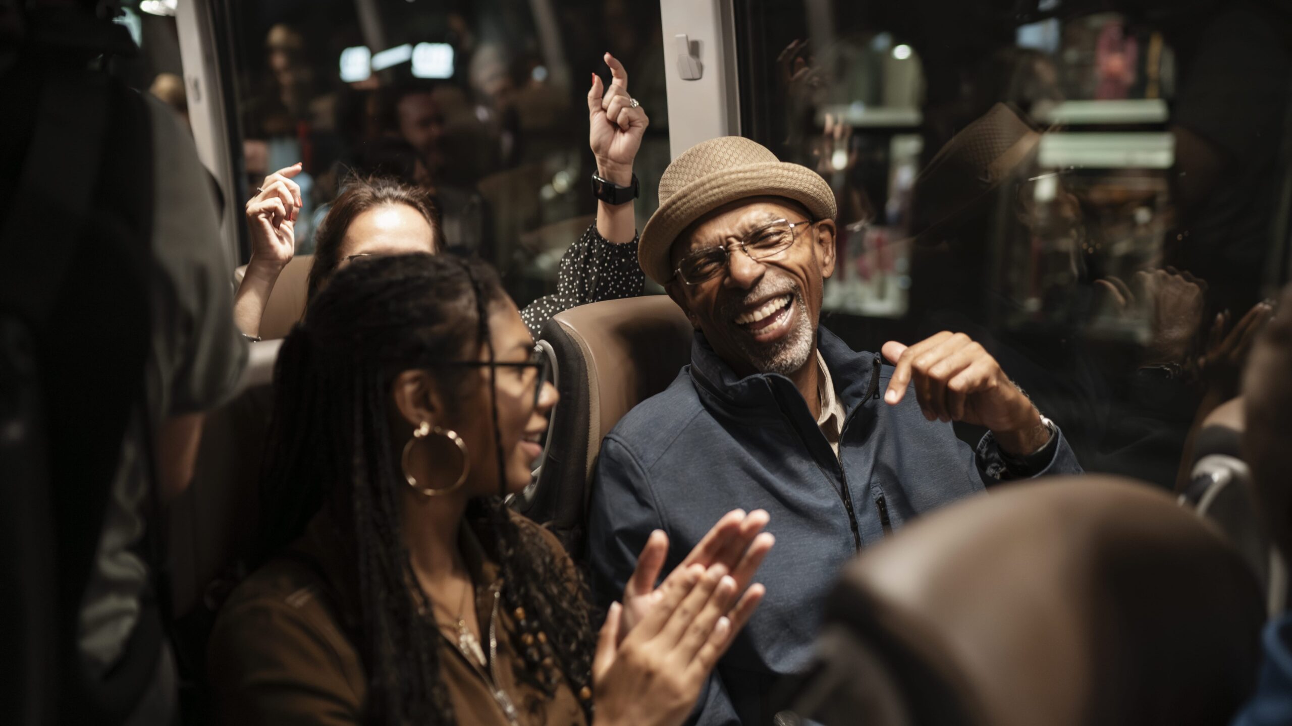 A joyful elderly man wearing a hat laughs heartily amidst a multi-generational group of people on a bus at night.