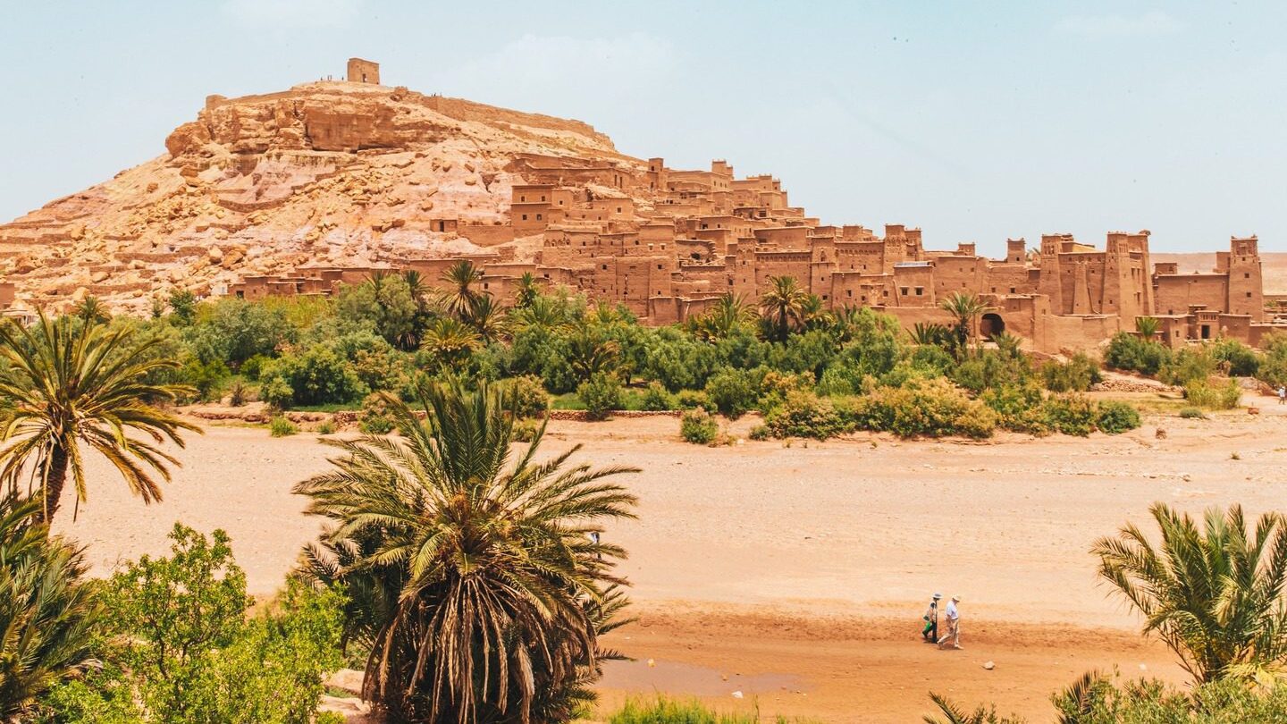 A landscape view of Ait Benhaddou, an ancient fortified village in Morocco, with a couple walking near a river and surrounded by lush palm trees during their Moroccan holiday.