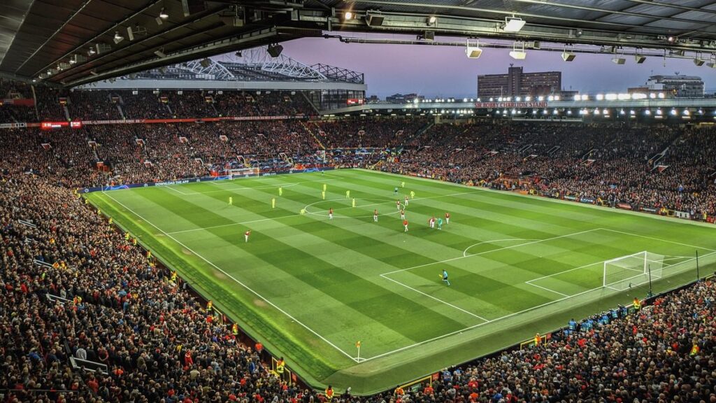 A night view of a packed football stadium during a game, showcasing a well-lit field, crowded stands, and an open roof.