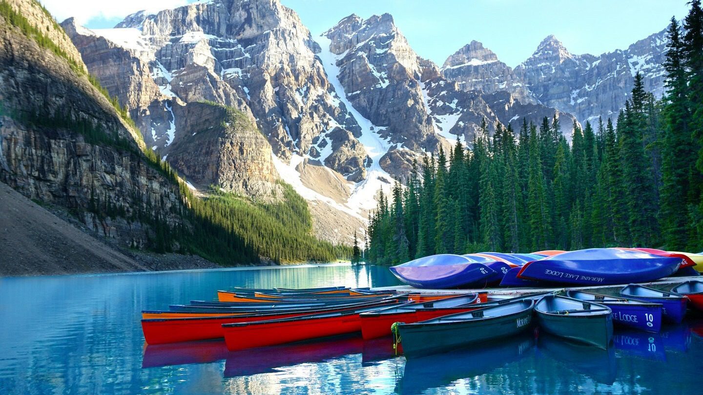 Canoes moored to a jetty on a blue lake surrounded by snowy peaks and pine forest