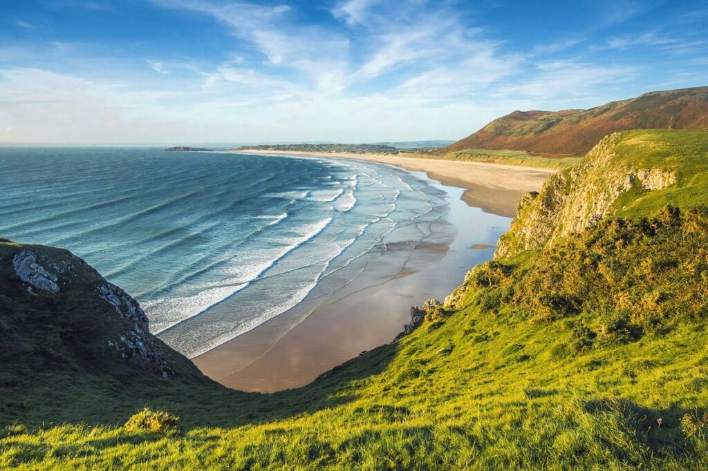 Scenic view of a sweeping coastline with sandy beach, gentle waves, and lush green hills under a blue sky during a tour to the UK.