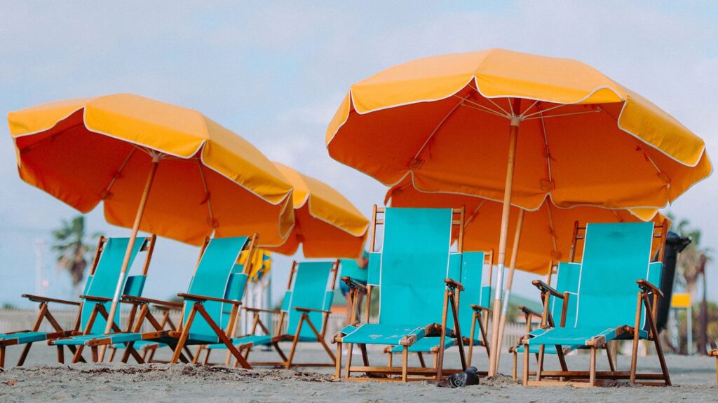 Row of teal beach chairs and orange umbrellas on sandy beach with palm trees in the background, perfect for planning your next holiday.