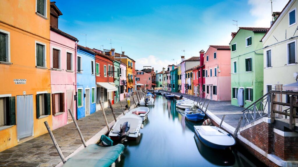 View looking down a narrow canal, lined by colorful houses on the island of Burano in Venice, Italy.