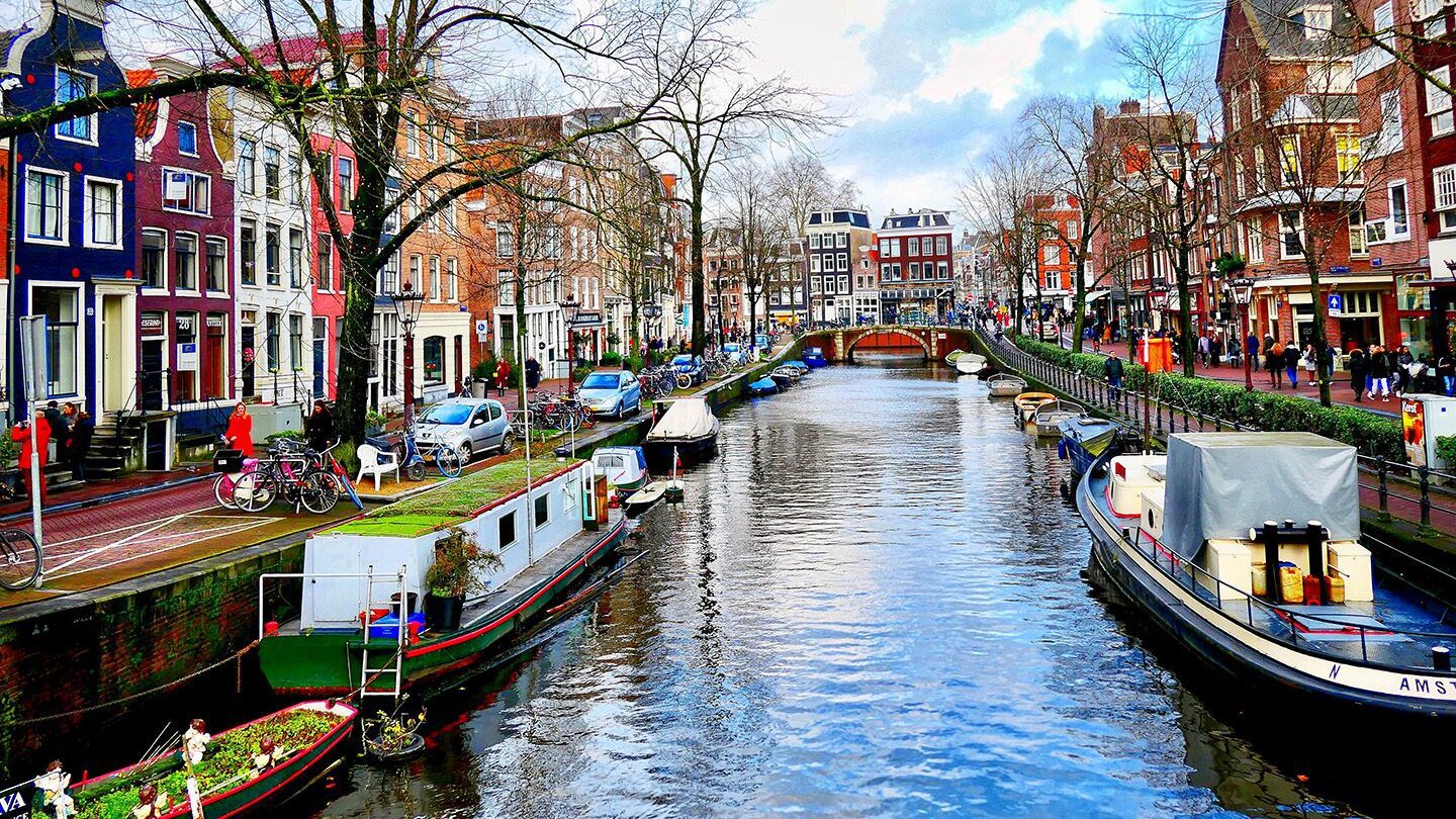 A vibrant scene of a canal in Amsterdam, one of the best places to live in the world, with colorful houses, moored boats, and people walking alongside the water.