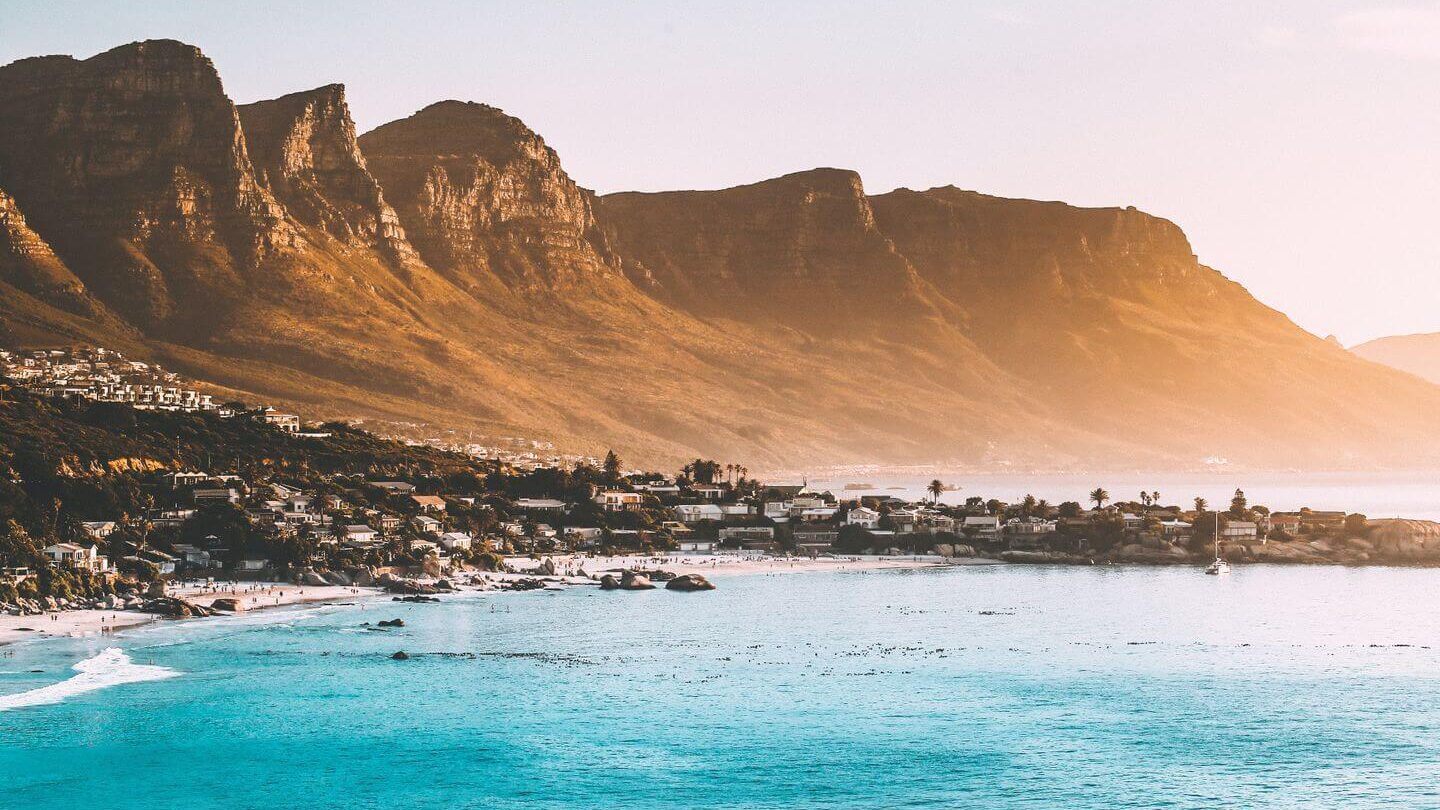 Coastal landscape at sunset with a clear blue sea in the foreground and a mountain range beside a town under a hazy sky, epitomizing why September is the best month for travel.