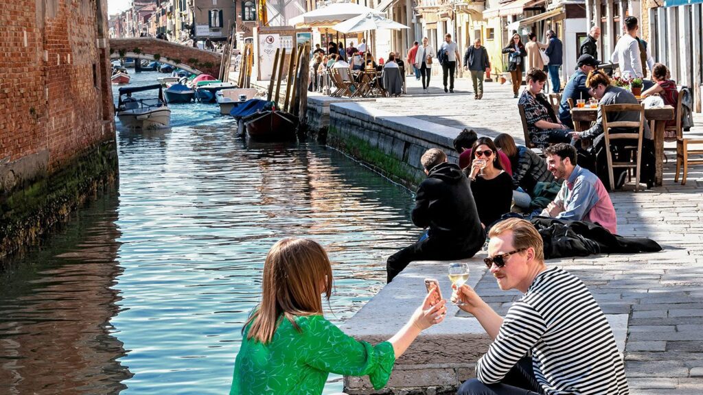 a man and woman pose with glasses of wine on the side of a canal in Venice
