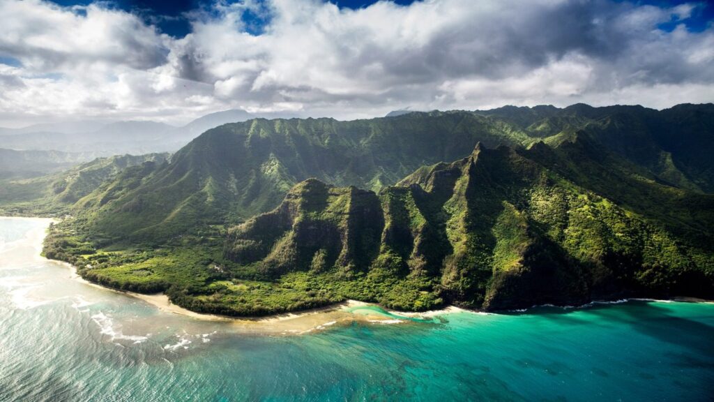 Aerial view of a lush, green mountain range beside a turquoise sea under a partly cloudy sky in one of the best places in Canada.