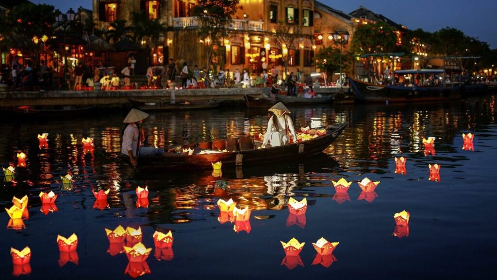 People wearing traditional Vietnamese conical hats in a small boat on a canal in Hoi An at night, placing paper lanterns on the water