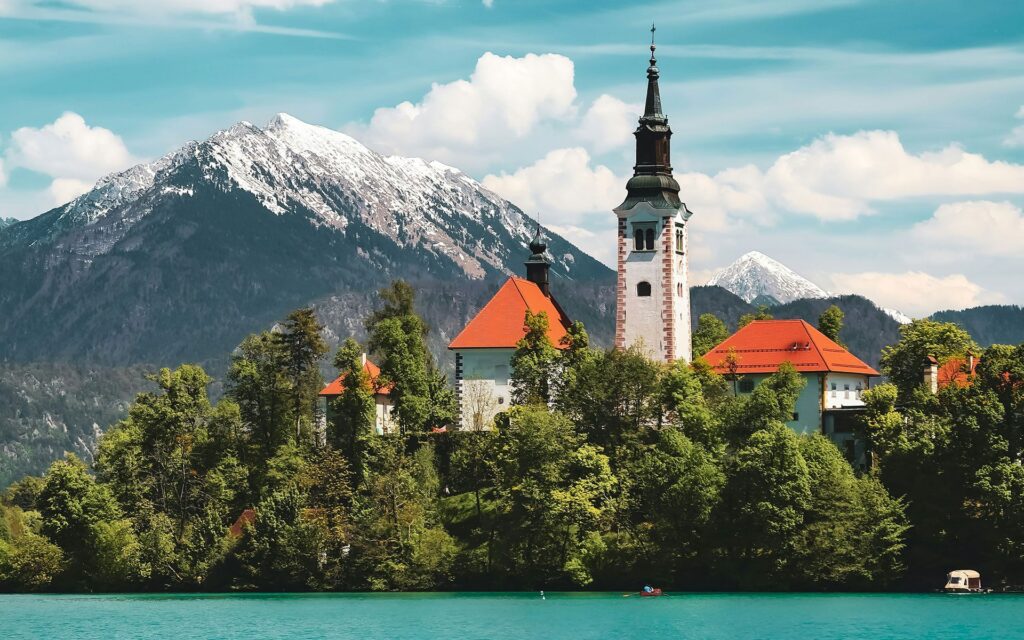 A church tower and red building roofs poke above the tree line of a lake island with a mountain in the background