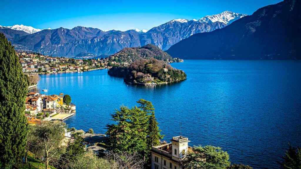 View of a serene lake surrounded by mountains in Italy, with a small town and green island visible under a clear blue sky in June.