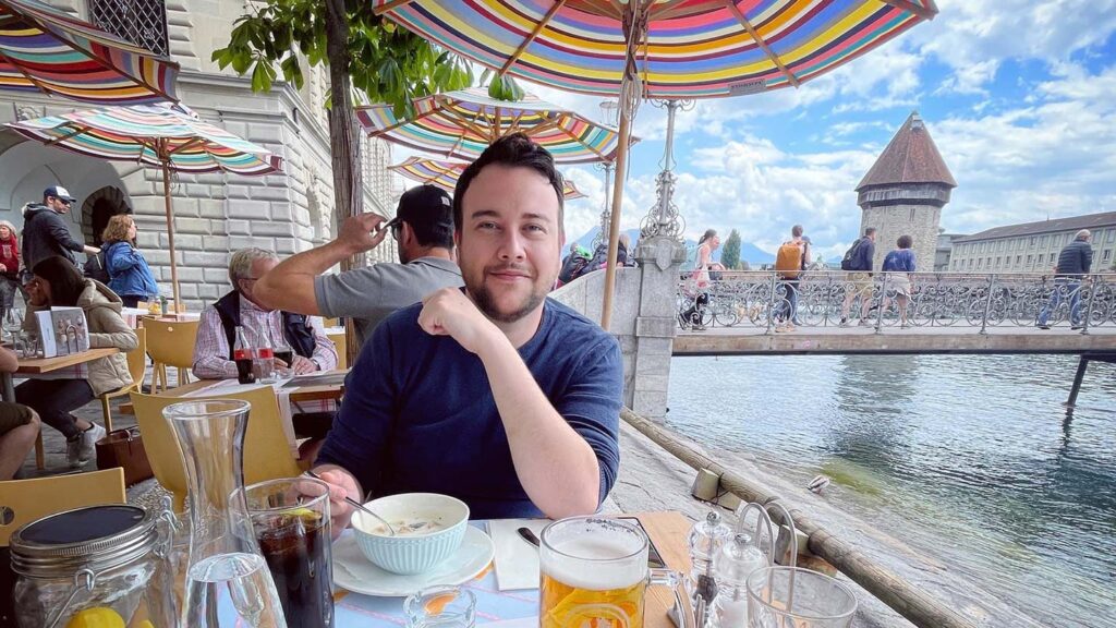 A man smiling at a riverside cafe under colorful umbrellas, with a historic tower in the background, possibly awaiting a proposal in Paris.